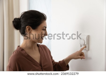 Smiling Indian woman using wall thermostat, smart home heating system, happy young female renter tenant homeowner setting comfortable temperature in apartment, energy saving and ventilation concept Royalty-Free Stock Photo #2369132279