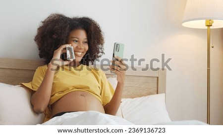 Smiling Black African American Pregnant woman holding showing ultrasound scan photos. Expecting mother having video call with family on smartphone at bed.