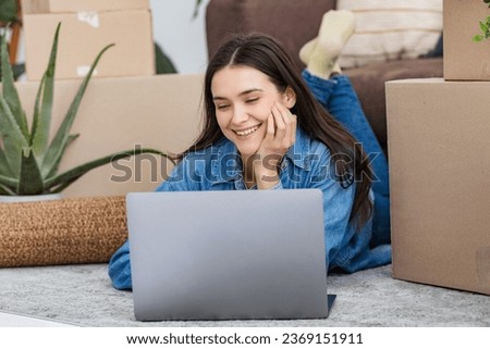Happy young woman moving her first new home while lying on the floor near the sofa among cardboard boxes, using laptop and smiling
