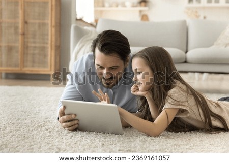 Caring young father spend time with adorable little preschooler daughter lying on floor together, look at digital tablet screen, make on-line purchase, watch cartoons, enjoy online internet content