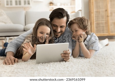Smiling dad spend time with little kids at home, friendly family lying together on floor in living room use digital tablet, choose funny videos, chat remotely with relatives, make on-line purchases