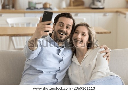 Beautiful millennial couple resting on sofa staring at cell phone screen, make selfie picture, calling to friends using videocall application spend weekend together at home. Leisure using modern tech