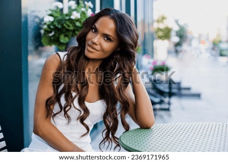 Beautiful portrait of an American girl with long curly hair sitting on a flyer in a cafe. woman smiling outdoors    Royalty-Free Stock Photo #2369171965