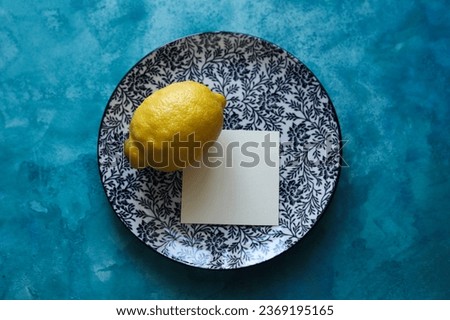 a still life with a plate with a floral pattern and a bright yellow lemon on a blue watercolor background