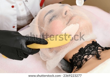 A beautician uses a spatula to apply cream to an already prepared face with gauze, a procedure for facial skin with couperosis. Royalty-Free Stock Photo #2369223277