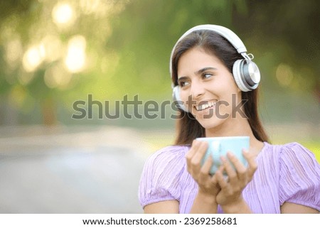 Happy woman wearing headphone laughing and listening music and drinking coffee in a park