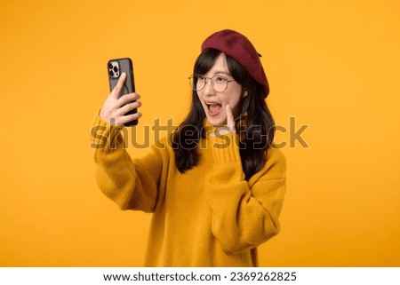 A joyful young woman wearing red beret and yellow sweater takes a selfie with her smartphone against a vibrant yellow background.