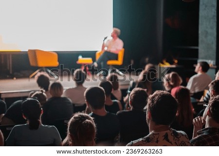 Crowd sitting in hall against blurred stage, while speaker showing presentation on big screen and speaking into mic during seminar Royalty-Free Stock Photo #2369263263