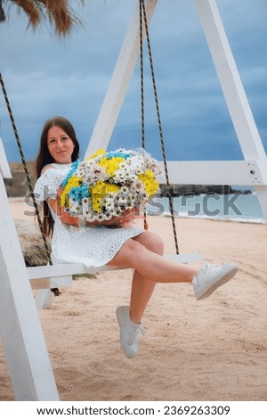 A beautiful girl with long hair in a white dress and a large bouquet of flowers, on a wooden swing on the seashore.