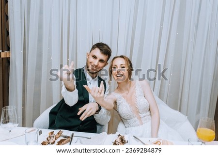A stylish smiling groom in a suit and a happy bride in a white dress sit at a table in a restaurant, cafe at a party and show the rings on their hands. Wedding photography, portrait.