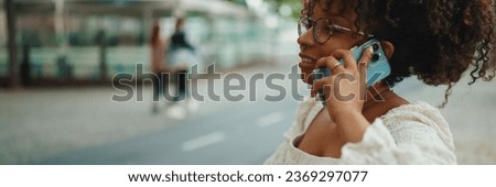 Young woman in glasses sits on a outdoor bench smiling and speaks on the smartphone. Positive woman using mobile phone outdoors in urban background.
