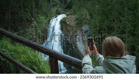 Woman takes pictures on the phone while traveling in Austria. Girl tourist films Golling waterfall in mountain forest.