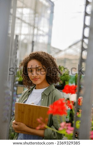 Smiling female gardener standing with clipboard on flower store background. Gardening concept