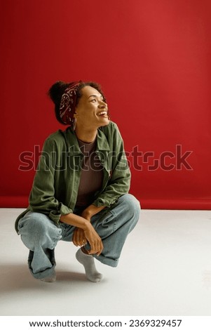 Cheerful stylish woman is squatting and posing with smile on red studio background and looks away