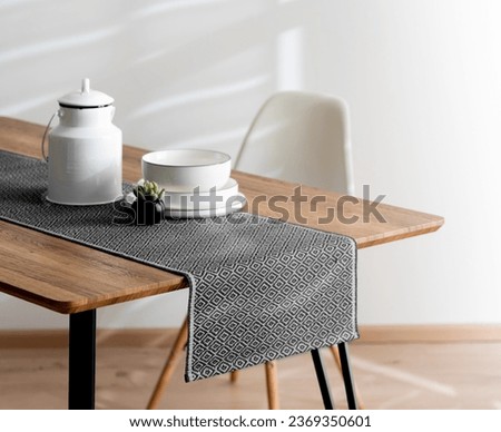 Modern Dining Room Featuring a Wooden Dining Table Adorned with a Grey Casual Linen Textured Table Runner, Teapot, and White Plates beside a White Plastic Dining Chairs, White Wall, Close Up. Royalty-Free Stock Photo #2369350601