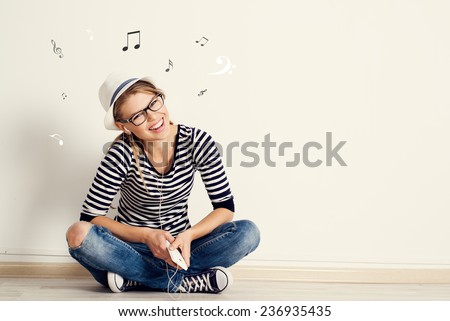 Portrait of happy female listening musical composition in earphones with sheet music and clef drawn on the wall. Young pretty Caucasian woman sitting on wooden floor in her house.  Royalty-Free Stock Photo #236935435