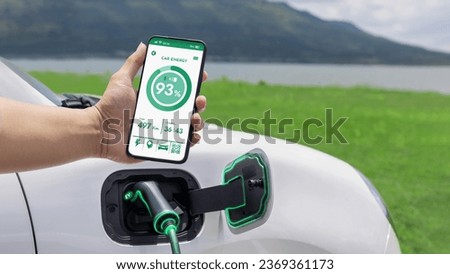 Businessman holding smartphone display battery status interface by smart EV mobile application while EV car recharging electricity from charging station with natural lake outdoor background.Peruse