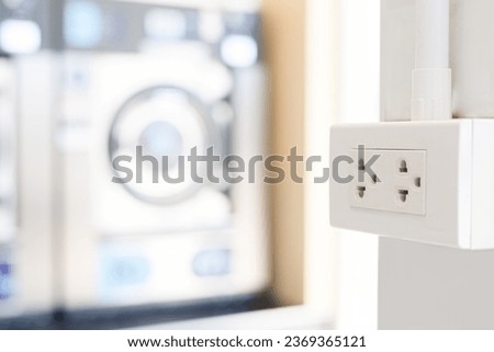 White plug on white wall with a lot of washing machine in a public laundromat in the background, plug socket, plug electric, plug outlet, backdrop. White outlet interior.
