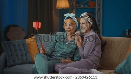 Medium-full photo capturing two young women wearing pajamas sitting on the couch and doing their evening skincare routine, taking photos using a selfie stick.