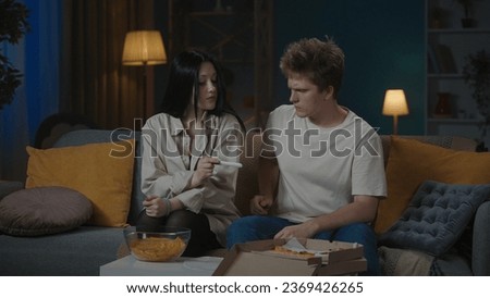 Portrait of teenage couple spending leisure time. Boy and girl sitting on the sofa, girl shows him positive pregnancy test, boy looks confused.