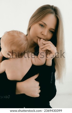 Young woman with baby in her arms. Blonde girl in black clothes smiling to your child on white background. Happy motherhood and breastfeeding concept. Photos with sun glare, soft focus, overexposure.