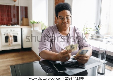 Sad frustrated plus size retired african american woman holding dollars in hands, counting her money, upset with low pension sitting at kitchen table next to window. Retired people and problems Royalty-Free Stock Photo #2369442599