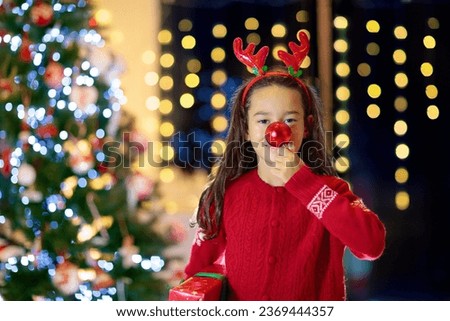 Child with reindeer nose and antlers on Christmas eve. Little girl holding a red bauble and Xmas present. Kids open Christmas gifts. Winter holiday fun. Family celebrating Christmas at home.