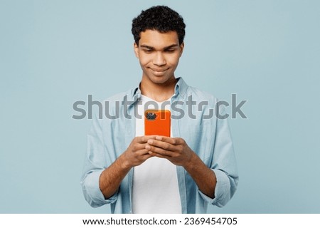 Young smiling happy cheerful fun man of African American ethnicity he wear shirt casual clothes hold in hand use mobile cell phone isolated on plain pastel light blue cyan background studio portrait