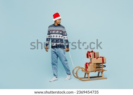 Full body young man wears knitted sweater Santa hat posing carry present box with gift ribbon bow on sled isolated on plain blue background. Happy New Year 2024 celebration Christmas holiday concept