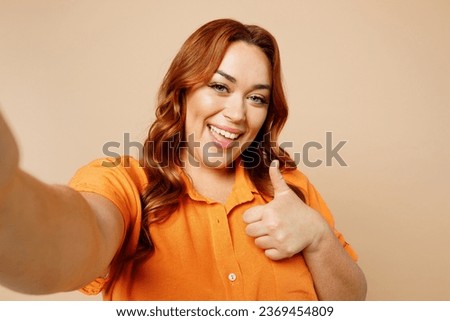 Close up young ginger chubby overweight woman wear orange shirt casual clothes do selfie shot pov mobile cell phone show thumb up isolated on plain beige background studio portrait. Lifestyle concept