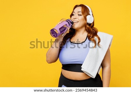 Young chubby overweight plus size big fat fit woman wears blue top warm up training listen to music in headphones drink water isolated on plain yellow background studio home gym. Workout sport concept