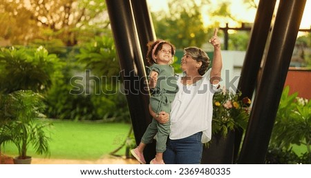 Happy Indian old grandmother holding cute little child girl in arms hands whirl dancing together outdoor home. Beautiful smiling woman sing song spinning small kid daughter enjoying summer holiday Royalty-Free Stock Photo #2369480335