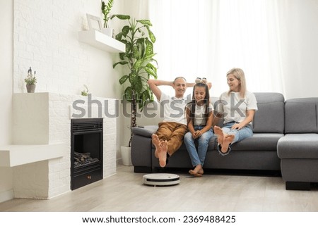 Young family resting on the couch while robotic vacuum cleaner doing its work at home. Household robots concept. Royalty-Free Stock Photo #2369488425