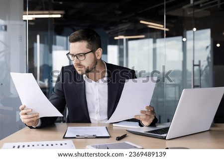 A young man office worker sits in the office at the table, holds papers in his hands, looks confusedly at documents and bills.