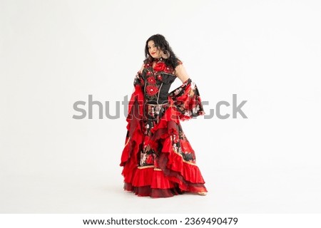 A young girl demonstrates a smart elegant red dress. Flamenco clothing. Gypsy romale style. Professional dancer isolated on white background. Royalty-Free Stock Photo #2369490479