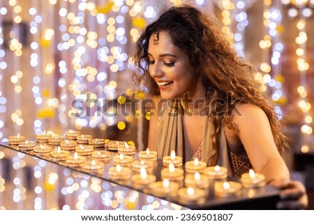 Beautiful woman smiling and holding diya. while standing against illuminated lights during diwali festival at home Royalty-Free Stock Photo #2369501807