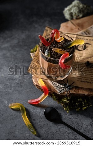 Sweets for Halloween party. Chocolate cream mousse in a glass. Chocolate puddings with chocolate cookie crumbs and gummy worms on dark festive background. Scary Halloween food, dessert. Recipe, menu