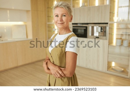 Portrait of happy aged elegant fashionable woman home owner with short haircut and smiling face posing against her new kitchen with folded arms, happy and proud with results of renovation Royalty-Free Stock Photo #2369523103