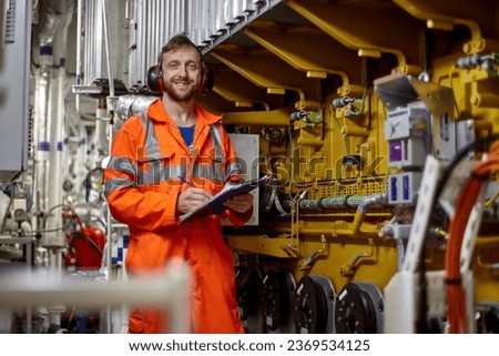 Portrait of young engineer with smile in orange coverall  doing check list in engine room. Royalty-Free Stock Photo #2369534125