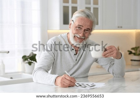 Senior man with cup of drink solving crossword at table in kitchen Royalty-Free Stock Photo #2369556653