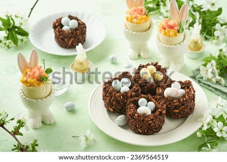 Easter chocolate nest cake with mini chocolate candy eggs with blossoming cherry or apple flowers on green background table. Creative recipe for Easter table with holiday decorations. Top view. Royalty-Free Stock Photo #2369564519