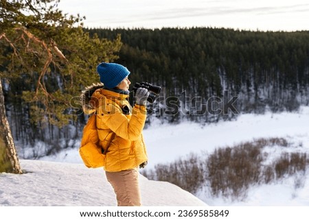 Young woman in yellow looking through binoculars at birds on snowy river against winter forest Birdwatching, zoology, ecology Research in nature, observation of animals Ornithology copy space Royalty-Free Stock Photo #2369585849