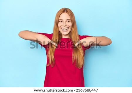 Redhead young woman on blue background surprised pointing with finger, smiling broadly.