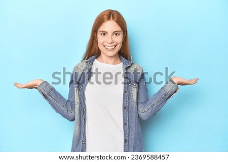 Redhead young woman on blue background makes scale with arms, feels happy and confident.