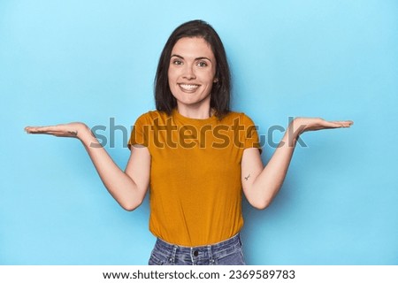 Young caucasian woman on blue backdrop makes scale with arms, feels happy and confident.
