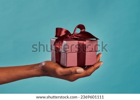 Close up shot of female hand holding a small gift wrapped with elegant brown ribbon isolated against blue background. Small craft Christmas gift in the hands of african american woman. 