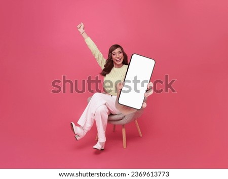 Portrait of happy Asian woman showing smartphone with white blank screen in hand sitting in armchair isolated on pink background.Gadget with empty free space for mockup, banner