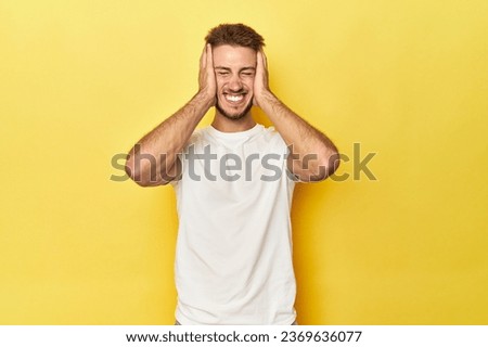 Young Caucasian man on a yellow studio background laughs joyfully keeping hands on head. Happiness concept.