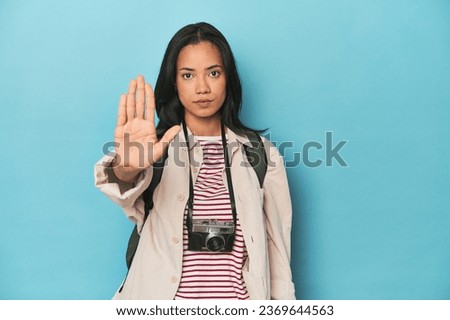 Filipina woman with camera and backpack on blue standing with outstretched hand showing stop sign, preventing you.