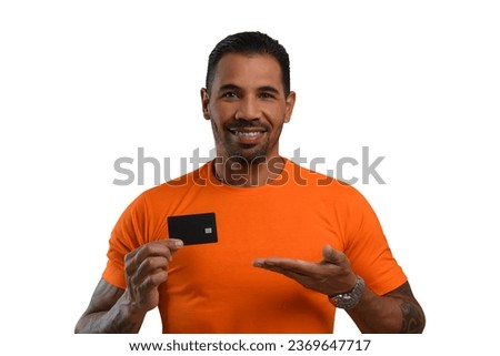 Man with a black credit card in his hand, pointing to the card, wearing an orange T-shirt, looking at the camera and smiling, white background